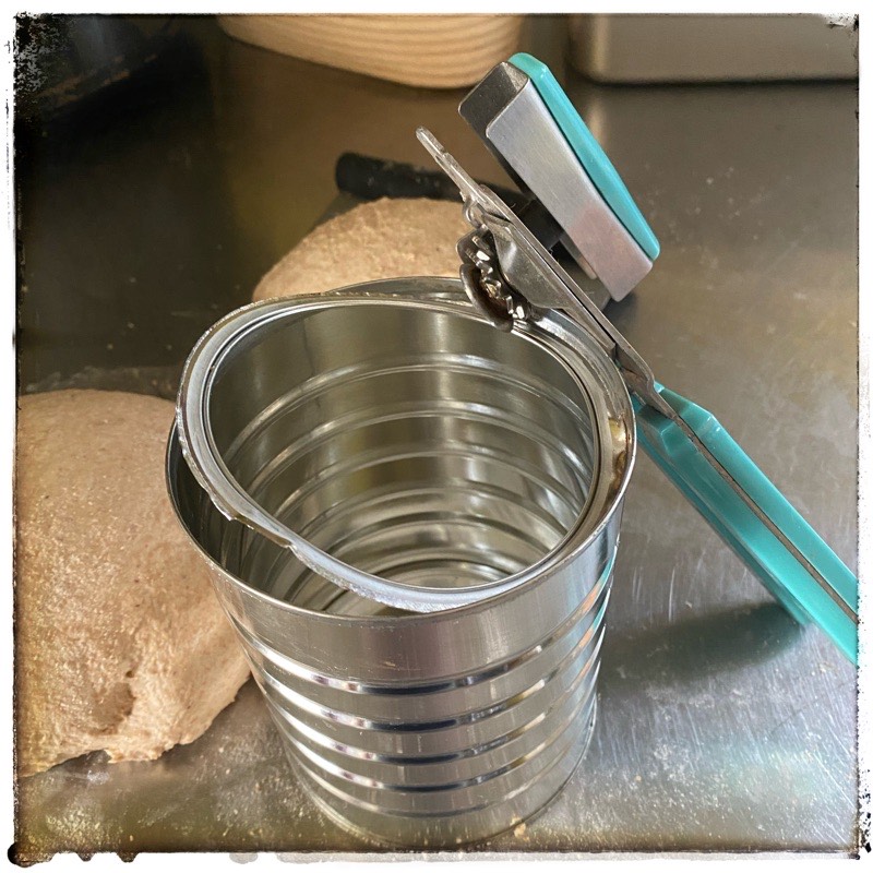 Coffee can showing removal of lip with aqua coloured can opener. Bread dough in the background. All on stainless steel counter. 